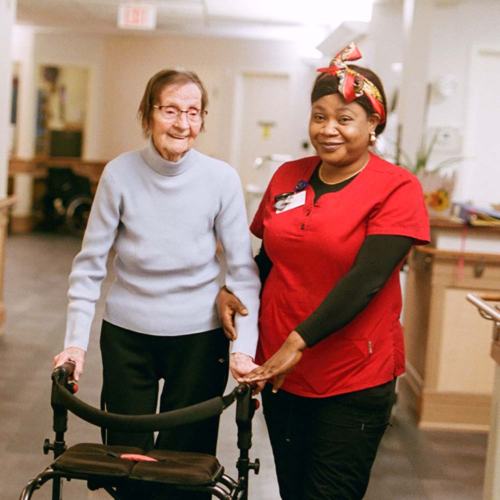 A female resident holding a walker stands beside a care worker in red scrubs.