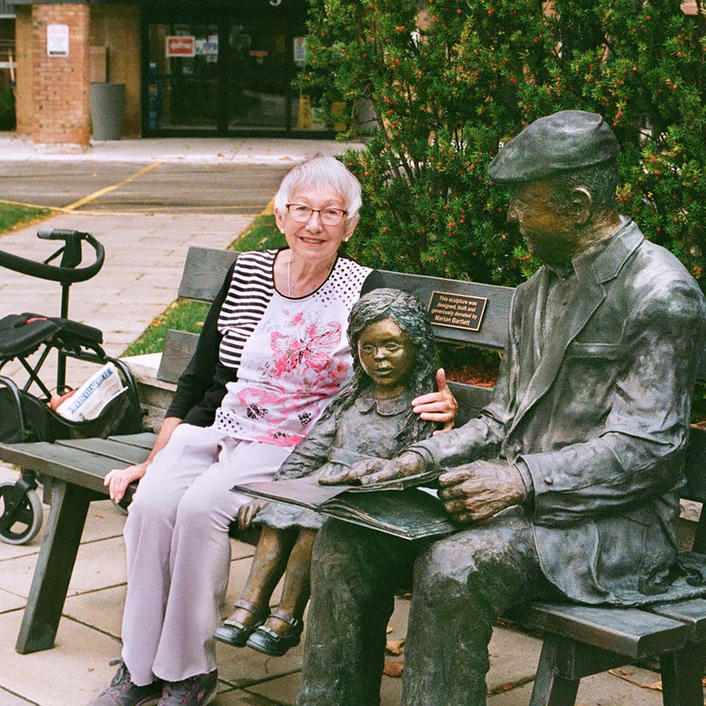 A woman with white hair and glasses sits on a bench beside a metal sculpture of a child and a man reading a book.