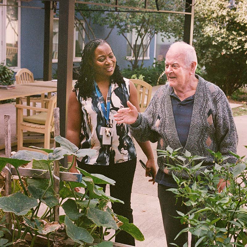 A senior man points to a plant in a garden, while a smiling care worker stands beside him