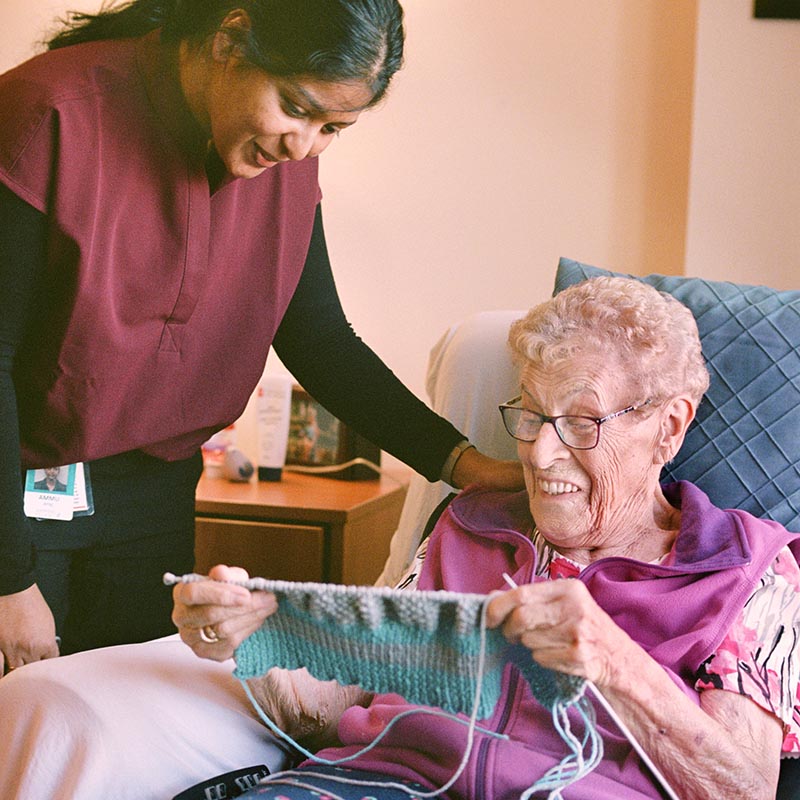 A senior woman shows a knitting project to a smiling care worker