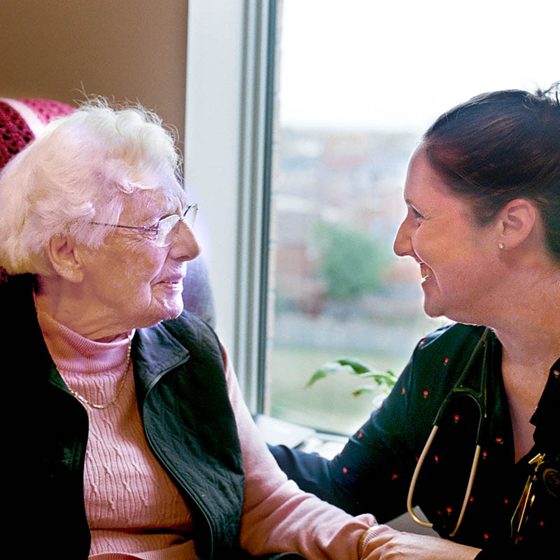 A smiling care worker speaks to a senior woman in a pink sweater