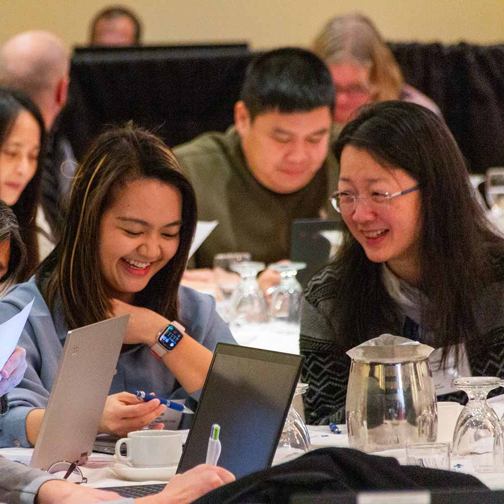 Two women seated at a table at a learning conference talk and laugh.