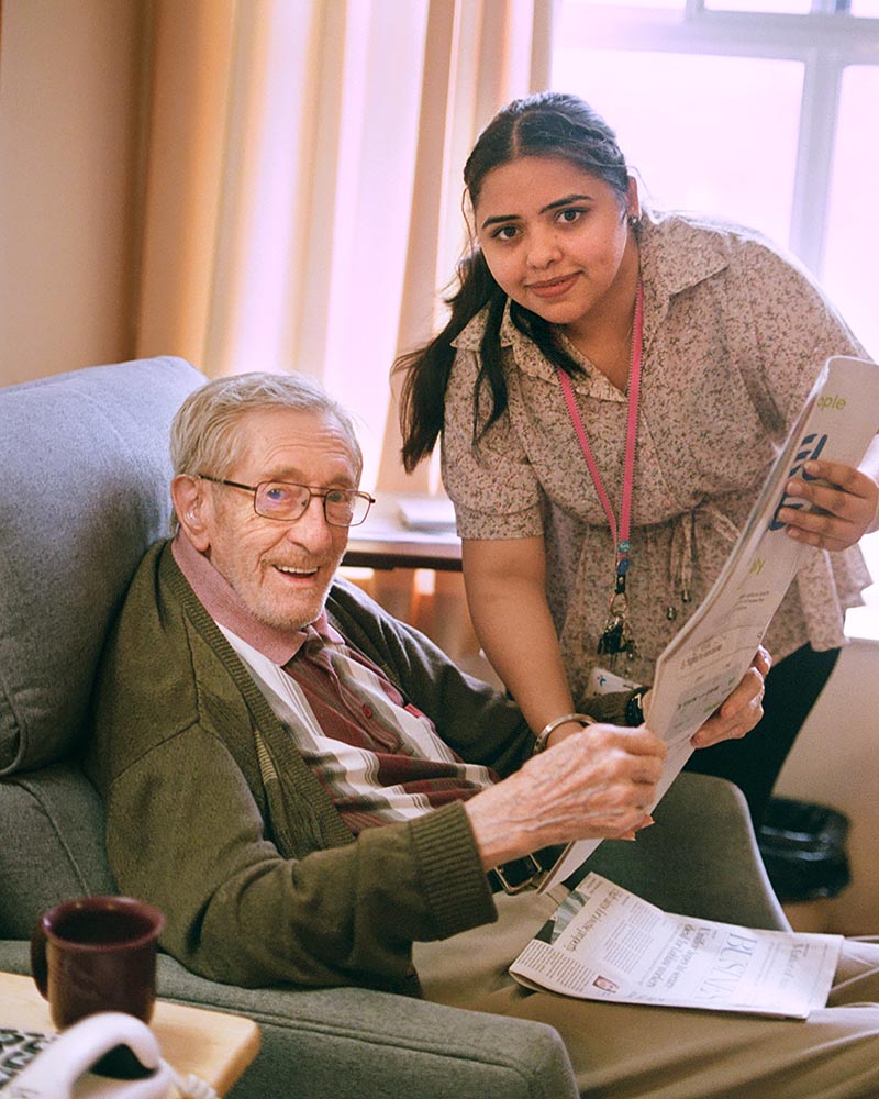 A senior man sits in an armchair and reads a newspaper with the assistance of a care worker