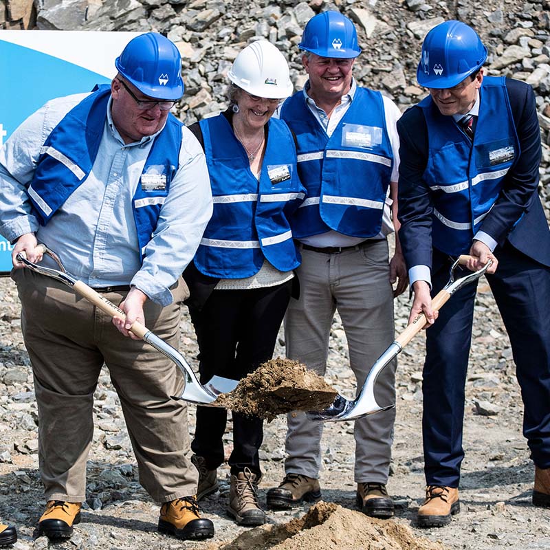 4 people wearing blue vests and hard hats smiling and shovelling dirt